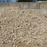 A large pile of crushed stone gravel is stacked outdoors in front of a concrete block wall, with bare trees and a clear sky visible in the background.
