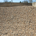 A large pile of small, light-colored stone gravel is shown outdoors, with a concrete wall and some trees visible in the background.