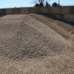 A large pile of loose gravel and stone sits in a concrete bin outdoors, with trees and a fence visible in the background under a clear sky.