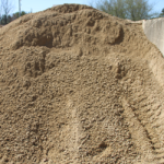 A large mound of loose, coarse sand or gravel with scattered pebbles and rough stone texture, possibly used for construction or landscaping. Trees and a concrete wall are visible in the background.