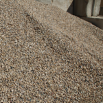A large pile of small, light-colored stone gravel is seen outdoors, next to a concrete wall under bright sunlight.
