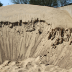 A large pile of sand with a deep, bowl-shaped indentation in the center, surrounded by uneven edges and ridges, with a few scattered stones beneath a clear sky.