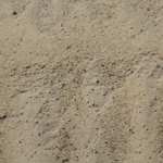 Fine, light brown sand with small pebbles and the occasional stone scattered across the surface. Wavy lines and gentle ridges show natural patterns created by wind or movement.