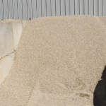 A large pile of small, light-colored stone gravel is contained by concrete walls, with a corrugated metal building in the background. Sunlight casts shadows on part of the gravel.
