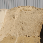 A large pile of light brown sand or fine gravel mixed with stone sits in front of a corrugated metal wall, with tire tracks visible in the sand at the bottom of the image.