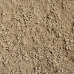 A close-up view of dry, loose, and light brown soil with small clumps, scattered stones, and a granular texture covering the entire frame.