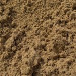 Close-up view of a pile of dry, loose sand with a rough, uneven surface, featuring small clumps and grains of sand visible throughout.